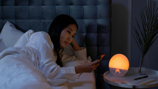 Woman using her phone in bed next to dim sunrise alarm clock